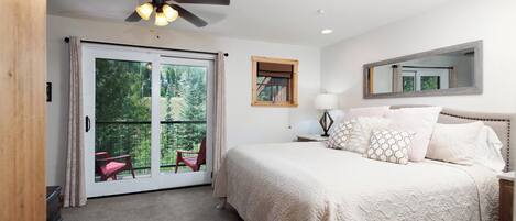 A bedroom with a bed, a ceiling fan, a window with a view of trees, a nightstand with a lamp, and a mirror on the wall.