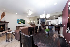 A spacious living room with a fireplace, a dining area with a long wooden table, and a kitchen with white cabinets and stainless steel appliances.