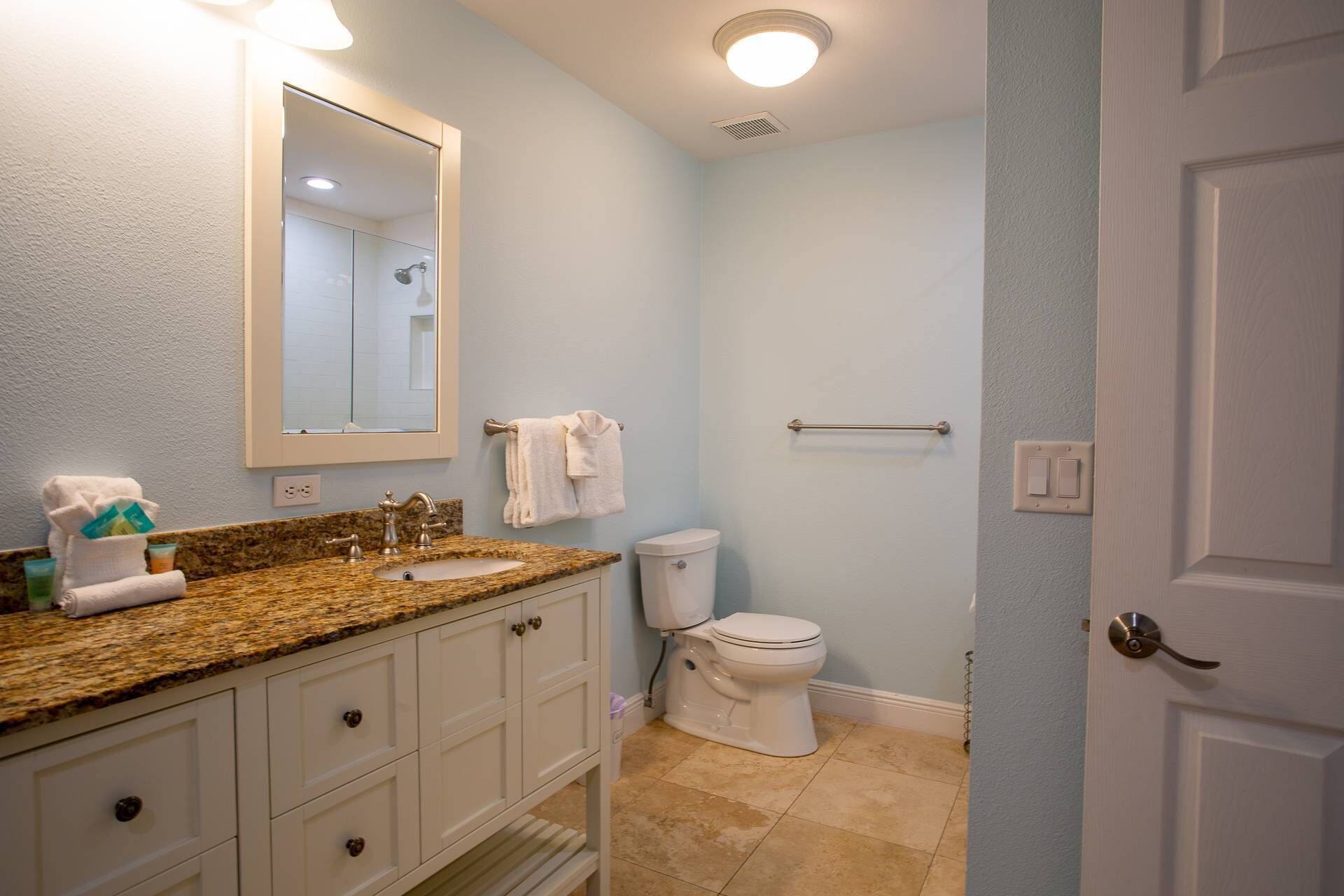 Un baño con lavamanos de encimera de granito, inodoro blanco, toallero y una puerta.