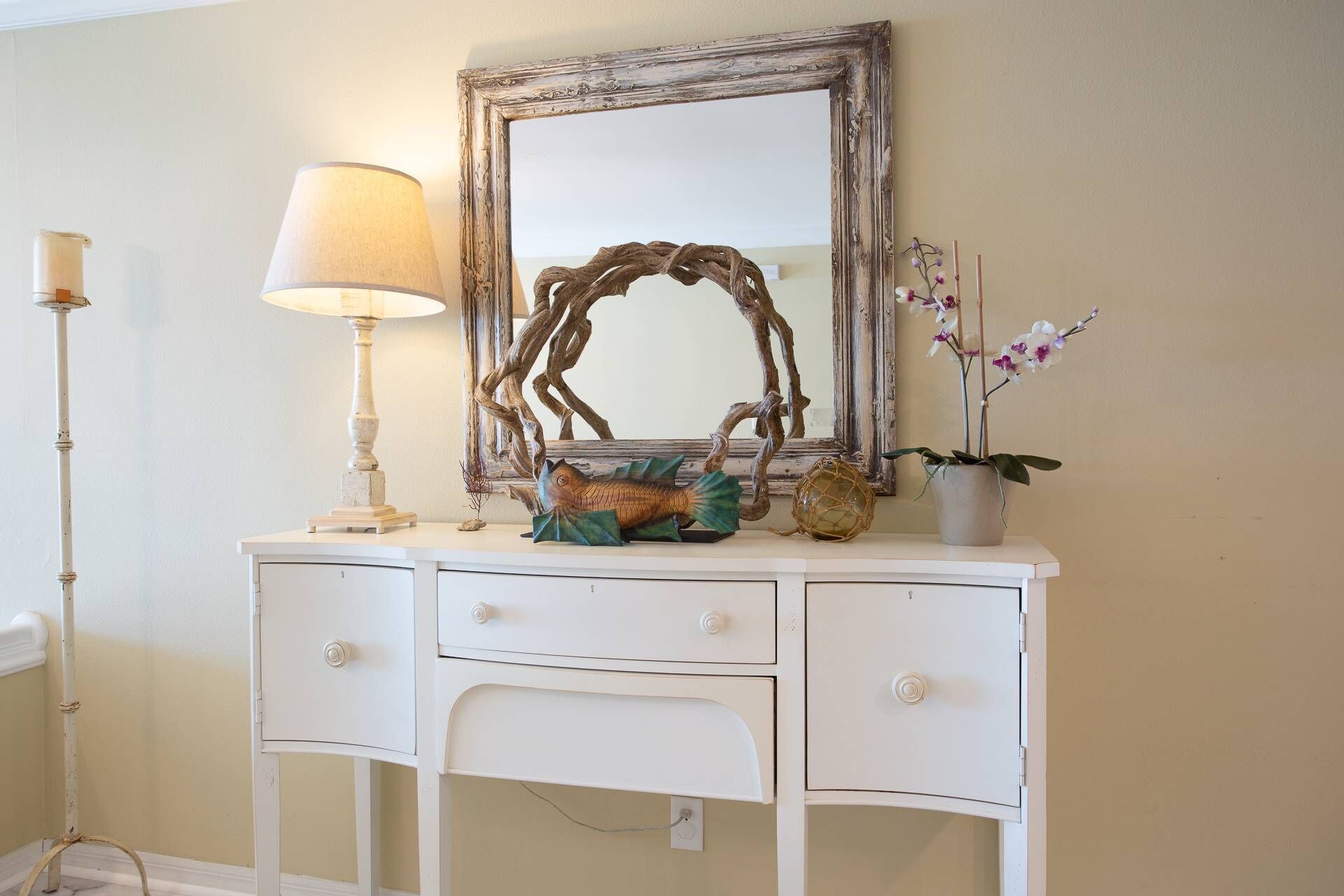 A white dresser with three drawers, a mirror, a lamp, decorative items, and a potted plant.