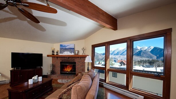 A living room with a fireplace, a large window with a mountain view, a television, and a ceiling fan. - Sitzmark townhome at Purgatory - Awesome views - Steps to slopes (Durango)