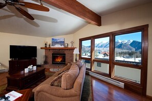 Un soggiorno con camino, una grande finestra vista montagna, televisore e ventilatore a soffitto.