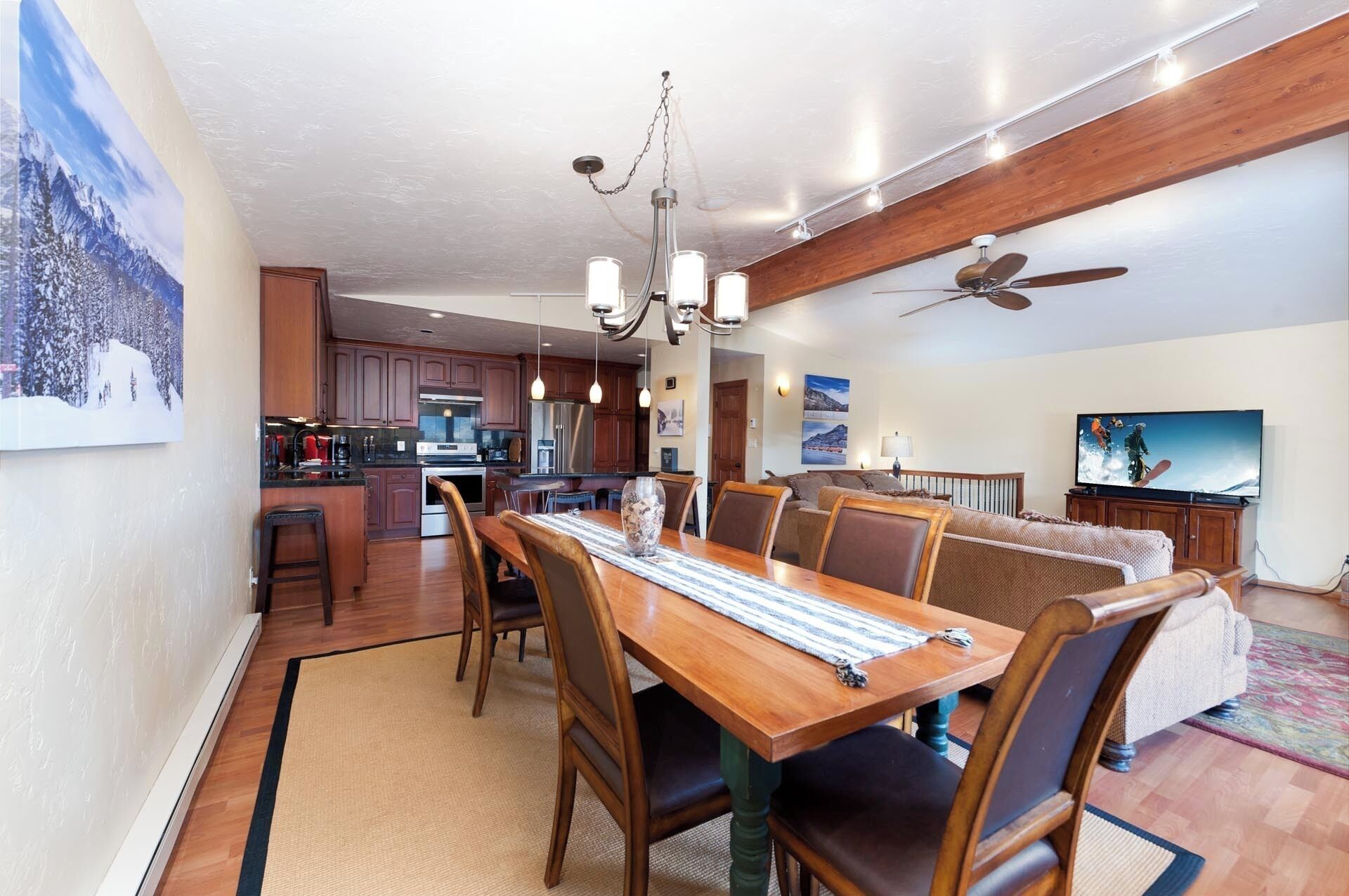 Un grand espace à manger avec une table et des chaises en bois, une cuisine à l’arrière-plan et un ventilateur de plafond.