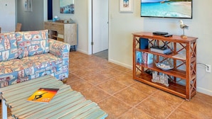 A living room with a patterned sofa, a wooden coffee table, a bookshelf, and a TV displaying a beach scene.