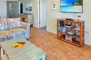 A living room with a patterned sofa, a wooden coffee table, a bookshelf, and a TV displaying a beach scene.