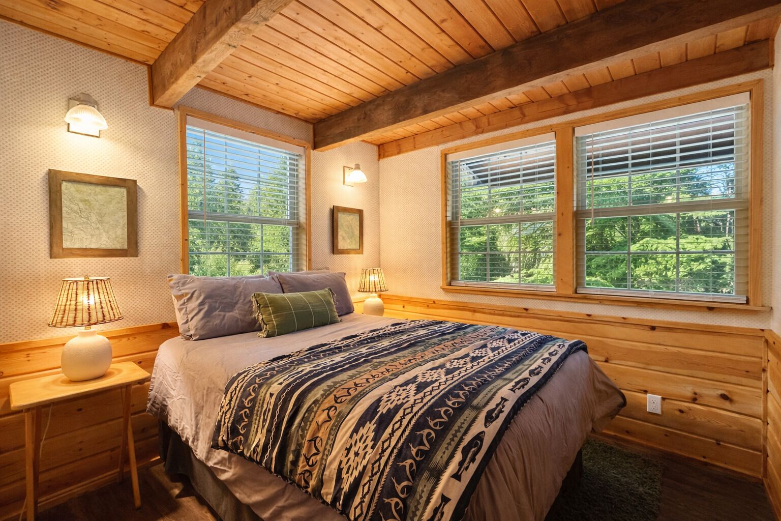 Une chambre confortable avec un plafond en bois, deux fenêtres équipées de stores, un lit avec une literie à motifs et des lampes de chevet.
