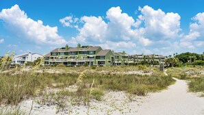Una proprietà sulla spiaggia con diversi edifici dai tetti verdi, un sentiero sabbioso che conduce alla spiaggia e un cielo azzurro limpido.