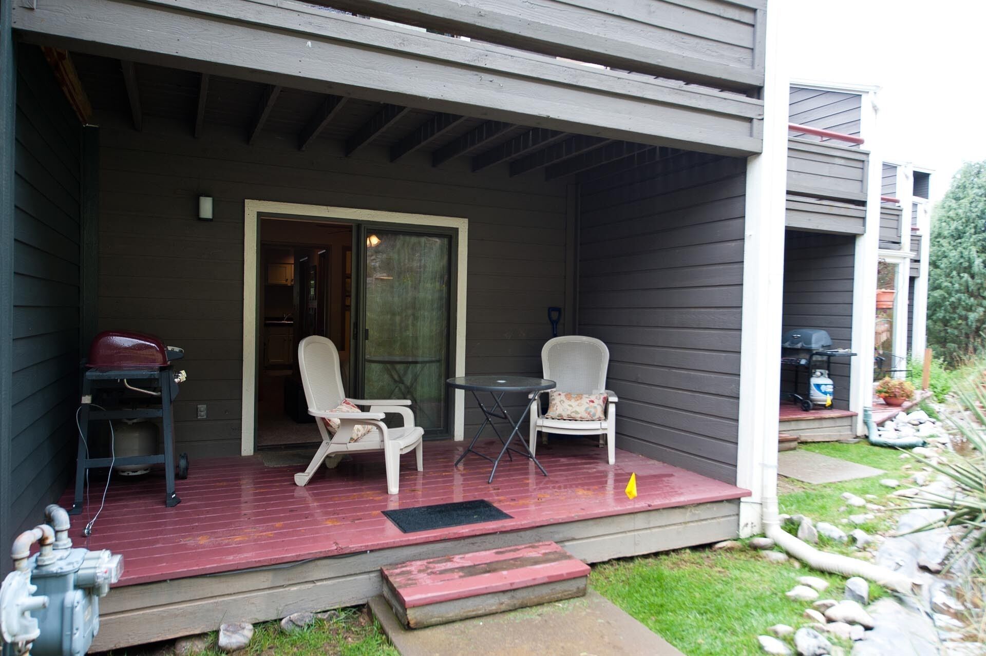A covered porch with two white chairs and a small table, a barbecue grill, and a gas tank outside a house with dark siding.