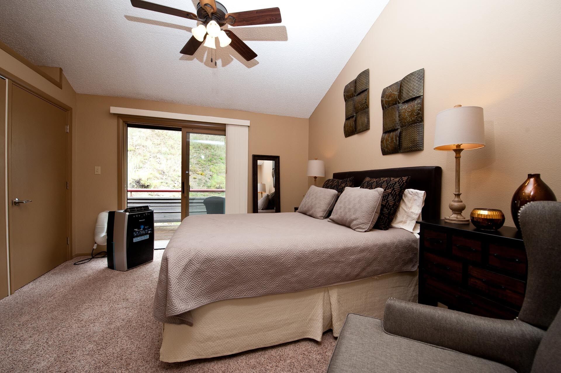 A bedroom with a bed, a desk, a chair, a ceiling fan, and a view of the outdoors.