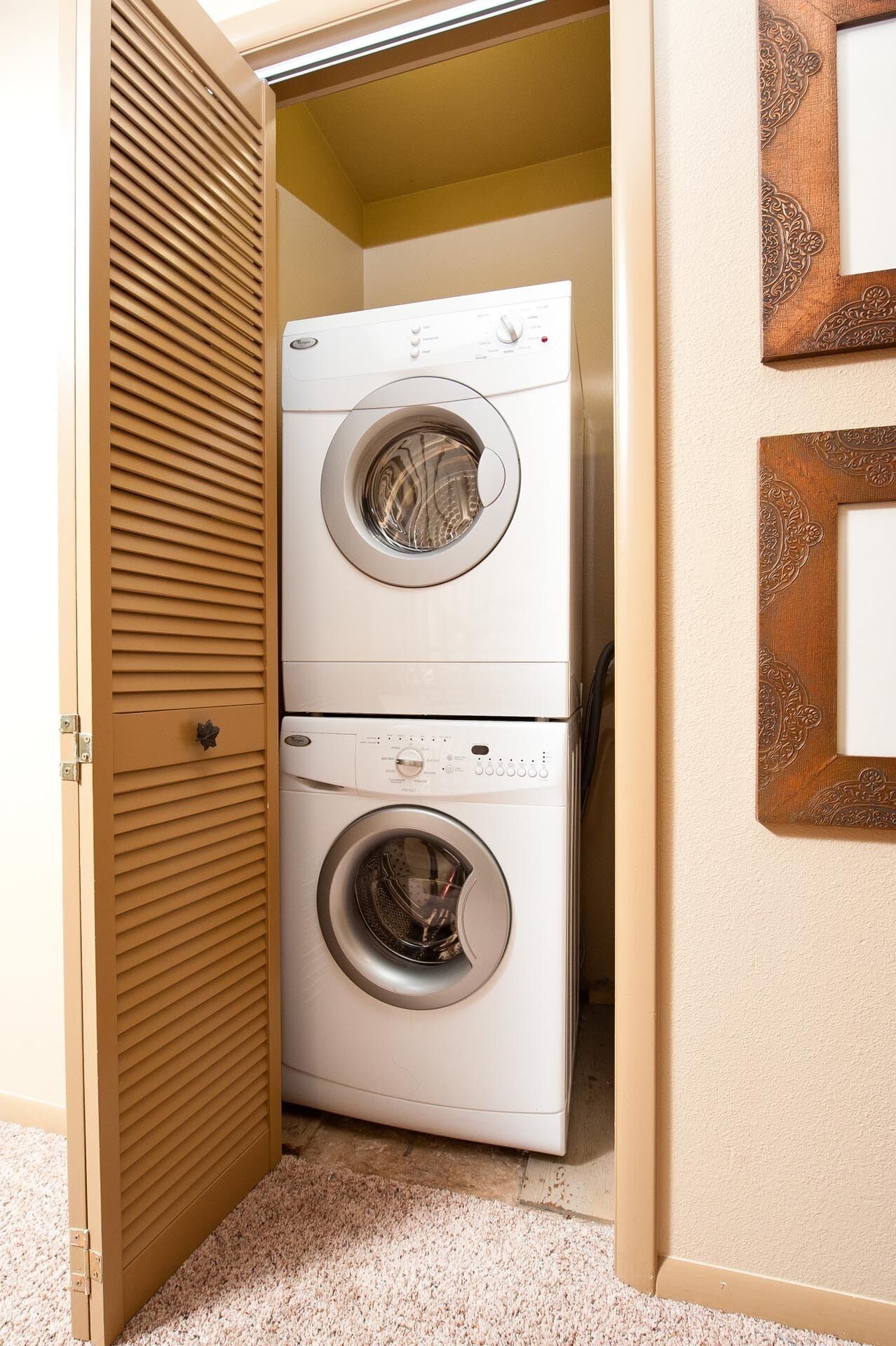 Un lavadero pequeño con dos lavadoras de carga frontal apiladas, una puerta de madera con manija y una alfombra color beige.