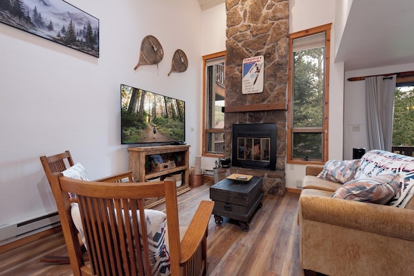 A living room with a stone fireplace, a television mounted on the wall, and a large window.