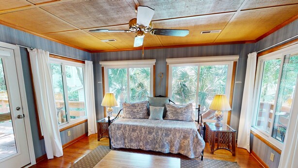 A spacious bedroom with a large bed, two bedside tables with lamps, a ceiling fan, and large windows offering a view of greenery outside.