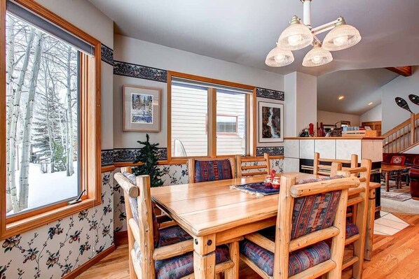 A dining area with a wooden table and chairs, large windows with a view of trees, and a kitchen in the background. - Village Townhome: Views, Location (Frisco)
