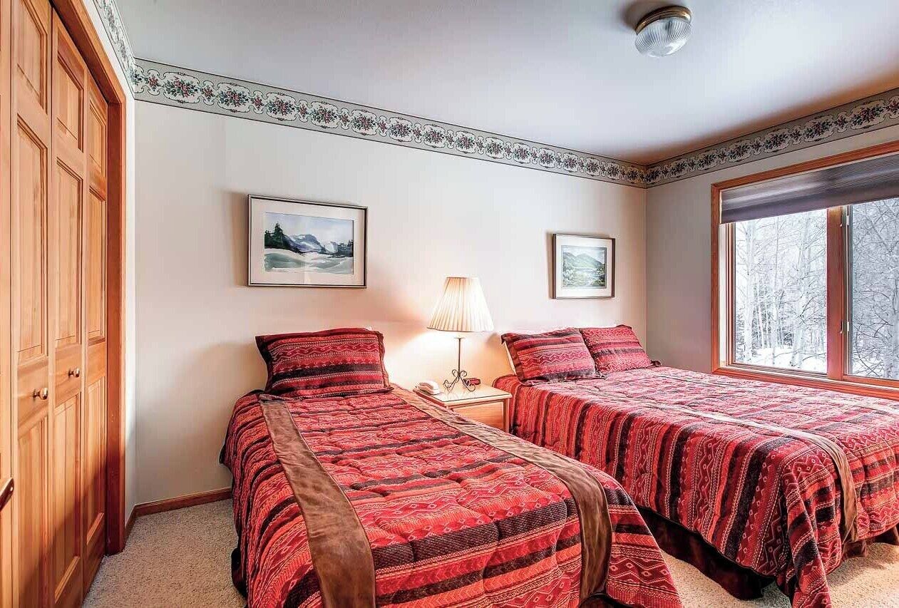 A bedroom with two beds, a wooden wardrobe, a nightstand with a lamp, and framed pictures on the wall.