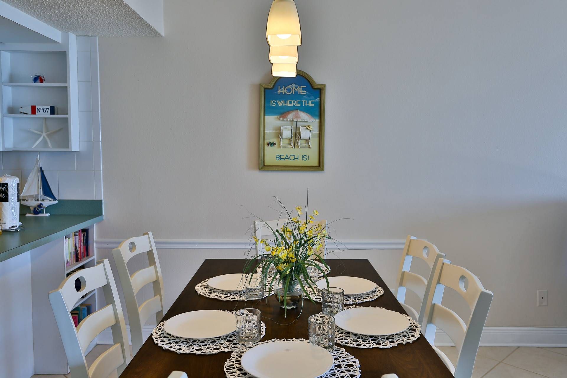 Un comedor con mesa de madera dispuesta para cuatro personas, rodeada de sillas blancas. Un tapiz decorativo sobre la mesa dice: 