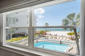 Blick auf einen Swimmingpool mit Liegestühlen und Palmen, einen Strand mit Sonnenschirmen und ein Gebäude mit weißer Fassade.