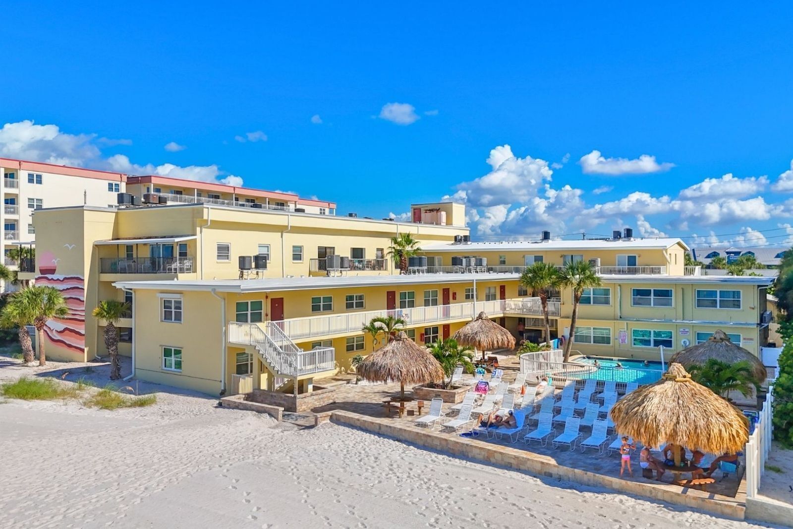 Un hôtel à étages, avec un espace piscine comprenant des chaises longues et des parasols, et une plage de sable en contrebas.