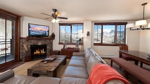 A cozy living room with a stone fireplace, a large flat-screen TV, a ceiling fan, and a view of snow-capped mountains through large windows. - Premier Building at Purgatory - Views - Ski in/Ski Out - Corner Unit (Durango)