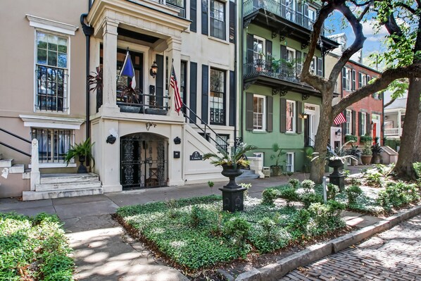 A row of historic buildings with a central entrance featuring a flag, steps, and a small garden area.