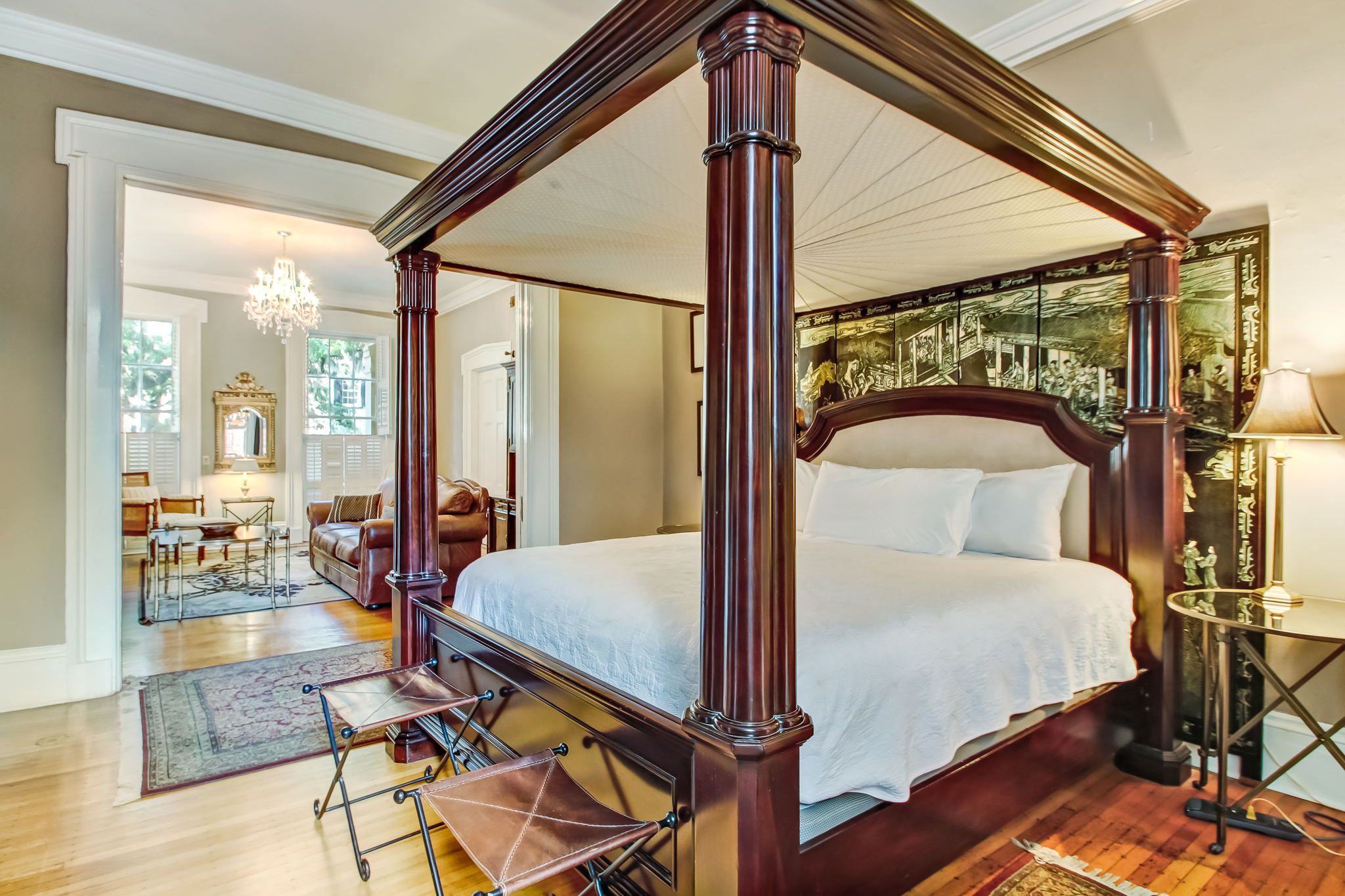 A four-poster bed with a dark finish, a chandelier, and a large mirror in a room with hardwood floors and a view of the outdoors.