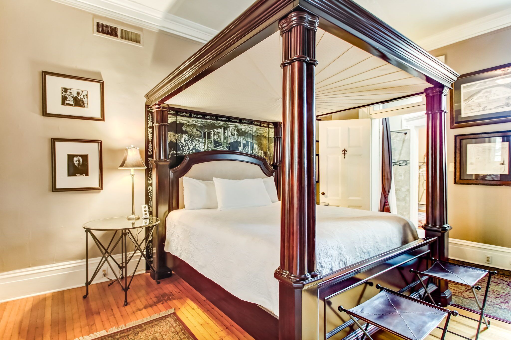 A four-poster bed with a dark finish, a small round table with a lamp, and framed pictures on the wall.