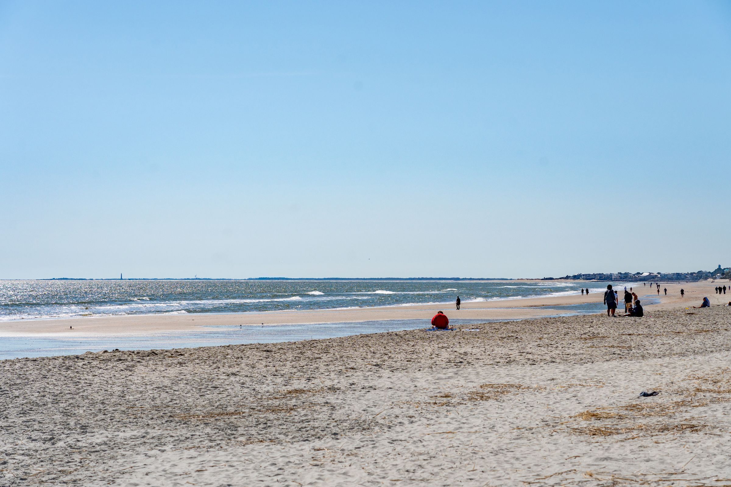 A beach with people walking and sitting, and a lighthouse in the distance.