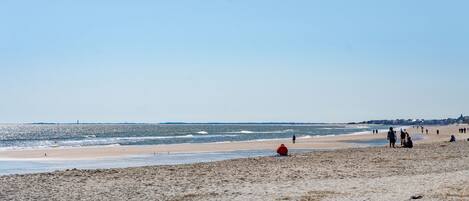 A beach with people walking and sitting, and a lighthouse in the distance.