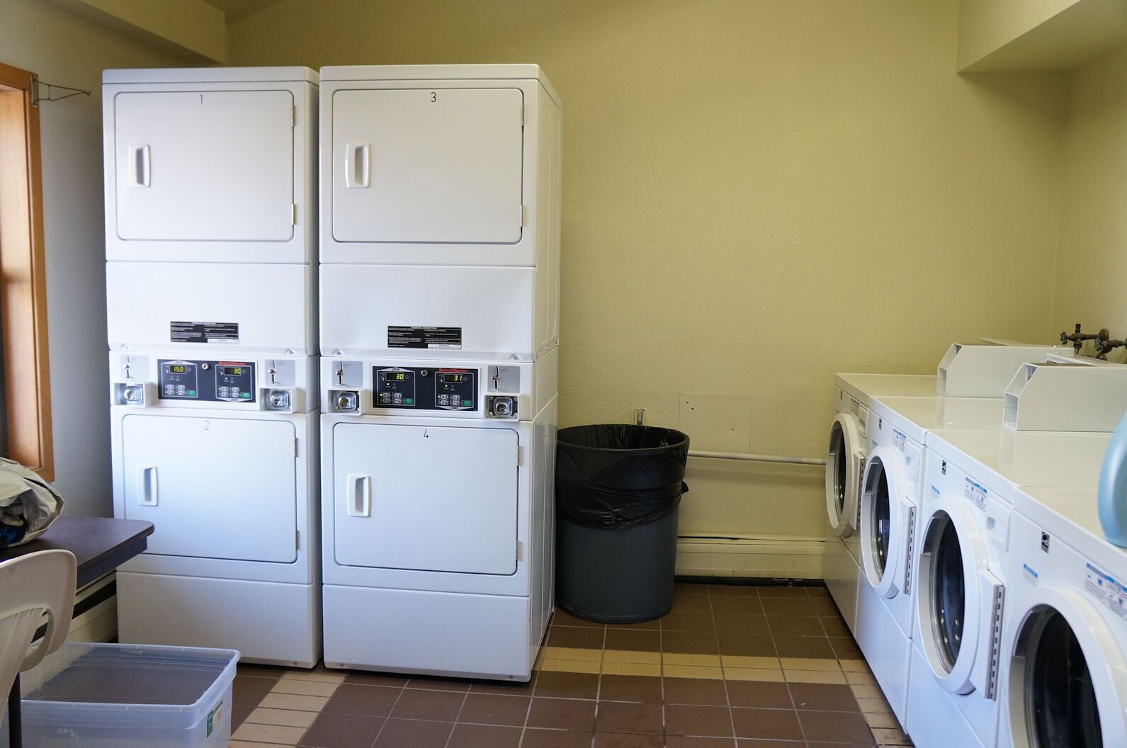 Une buanderie équipée de plusieurs sèche-linge superposés, d’une machine à laver et d’une poubelle.