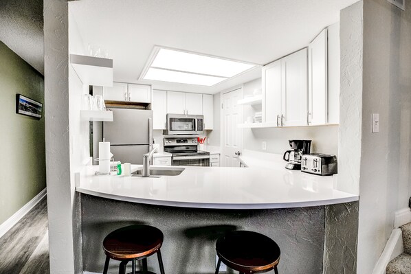 Una cocina moderna con armarios blancos, una isla central con taburetes, microondas, horno y cafetera.