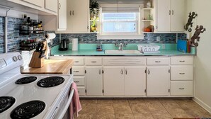 Une cuisine avec des meubles blancs, une cuisinière, un évier et une fenêtre avec des stores.