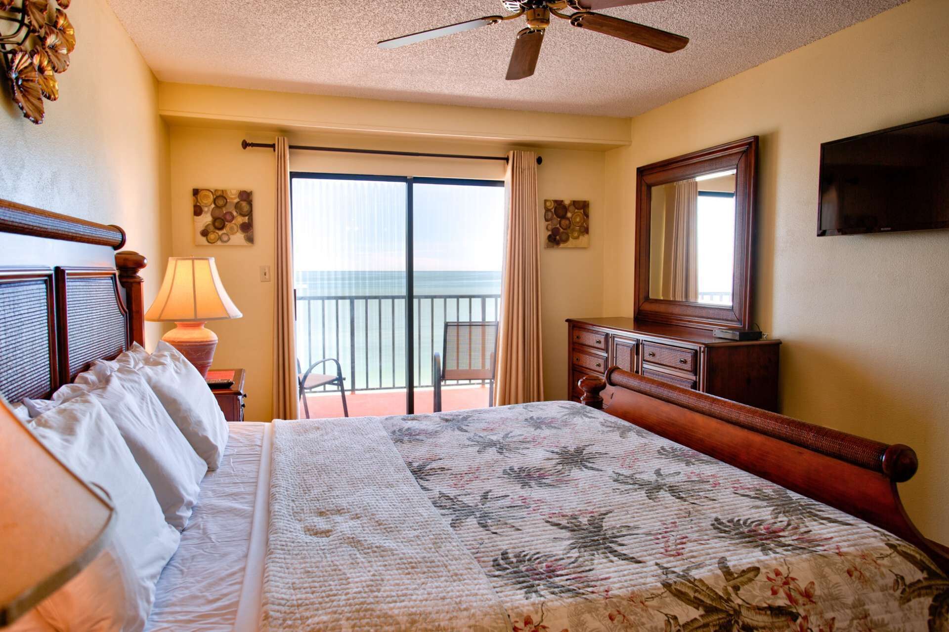 A hotel room with a bed, a ceiling fan, a balcony with a view, and a TV on the wall.
