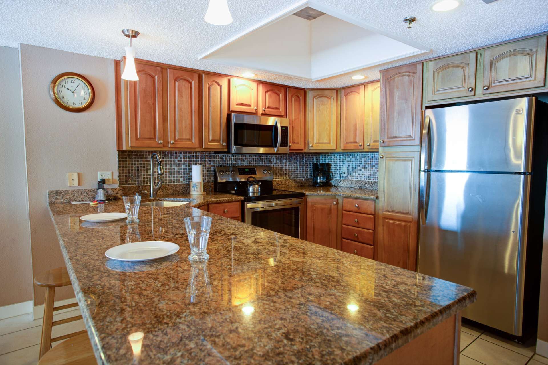 A modern kitchen with granite countertops, wooden cabinets, stainless steel appliances, and a clock on the wall.