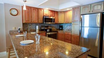 A modern kitchen with granite countertops, wooden cabinets, stainless steel appliances, and a clock on the wall.