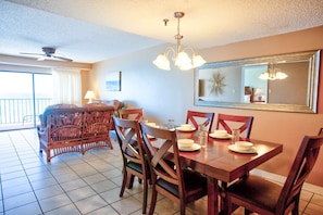 A dining area with a wooden table set for four, surrounded by chairs. There is a ceiling fan above and a chandelier hanging from the ceiling.