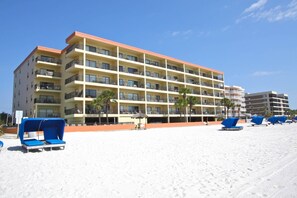 A multi-story hotel with balconies, a sandy beach in front, and blue lounge chairs.