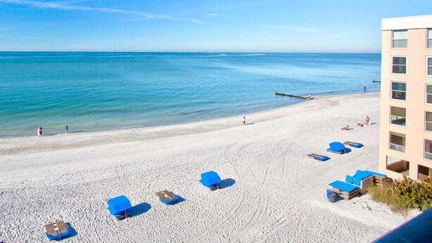 A beach with lounge chairs and a building in the background. - Beachfront Condo With Private Balcony Overlooking the Sand! Relax on the Beach or Lounge in the Pool (Madeira Beach)