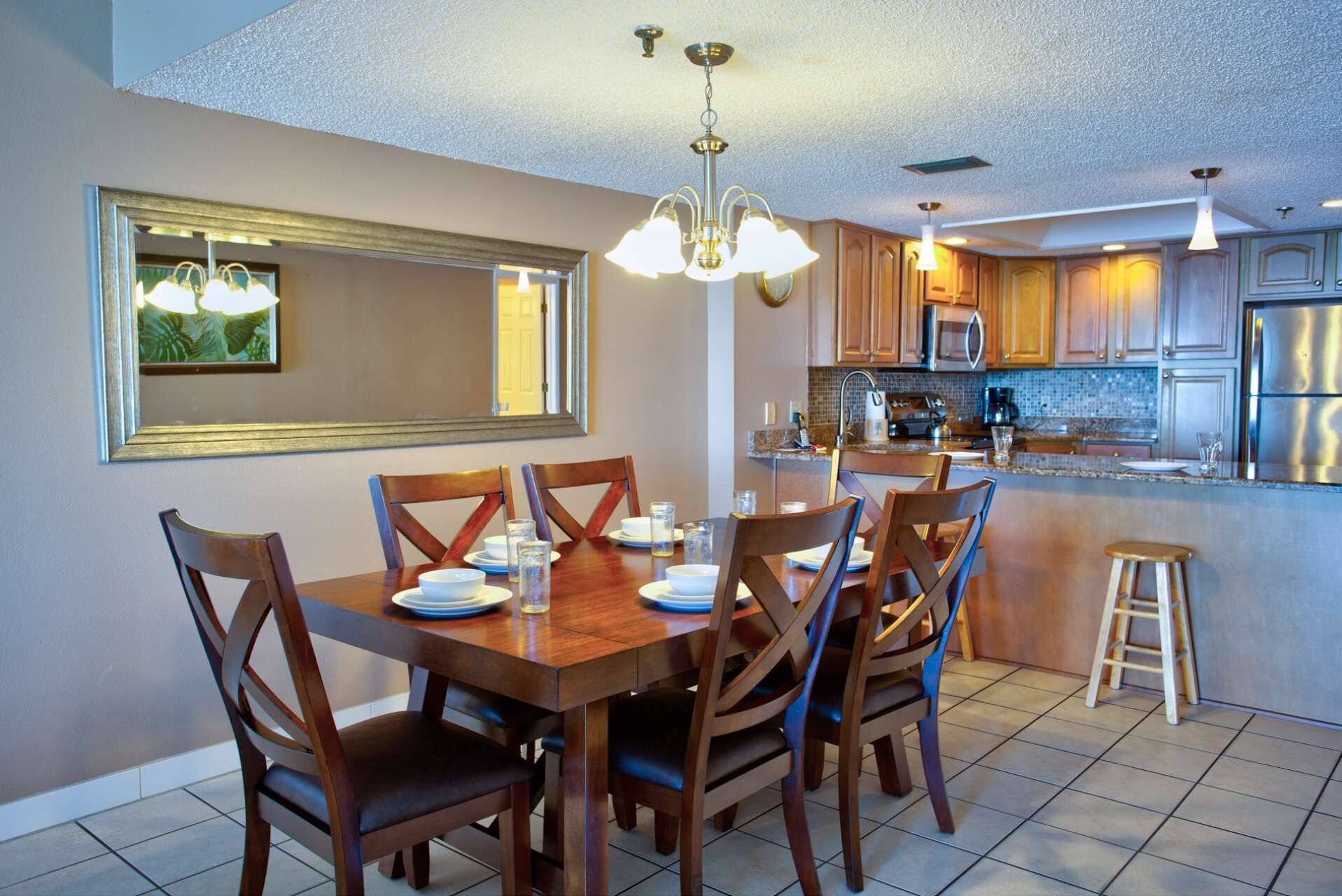 Un comedor con mesa y sillas de madera, una araña y una cocina con electrodomésticos modernos y salpicadero de azulejos.