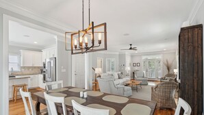 A spacious living room with a large wooden dining table, white chairs, and a modern chandelier.