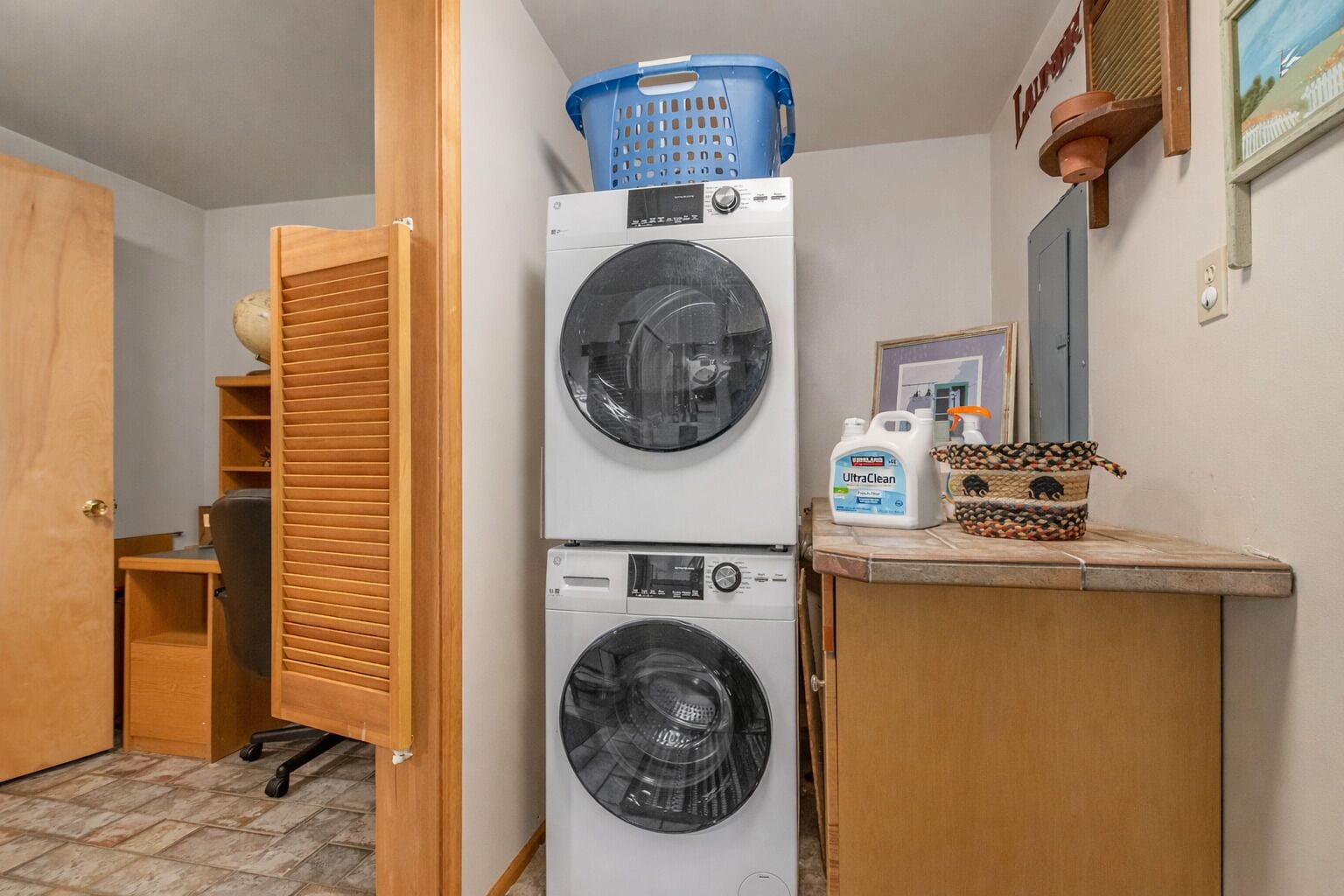 A laundry area with two stacked washing machines, a blue laundry basket on top, a bottle of laundry detergent, and a woven basket with a pig design.