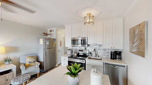A modern kitchen with stainless steel appliances, a white countertop, and a dining area with a striped chair and a small table. - Charming Oceanfront Condo with Balcony and Gorgeous Ocean Views! (Isle of Palms)