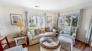 A well-lit living room with a beige sofa, a round coffee table, and a grey armchair. There are two windows with curtains, a lamp on a side table, and framed pictures on the wall.