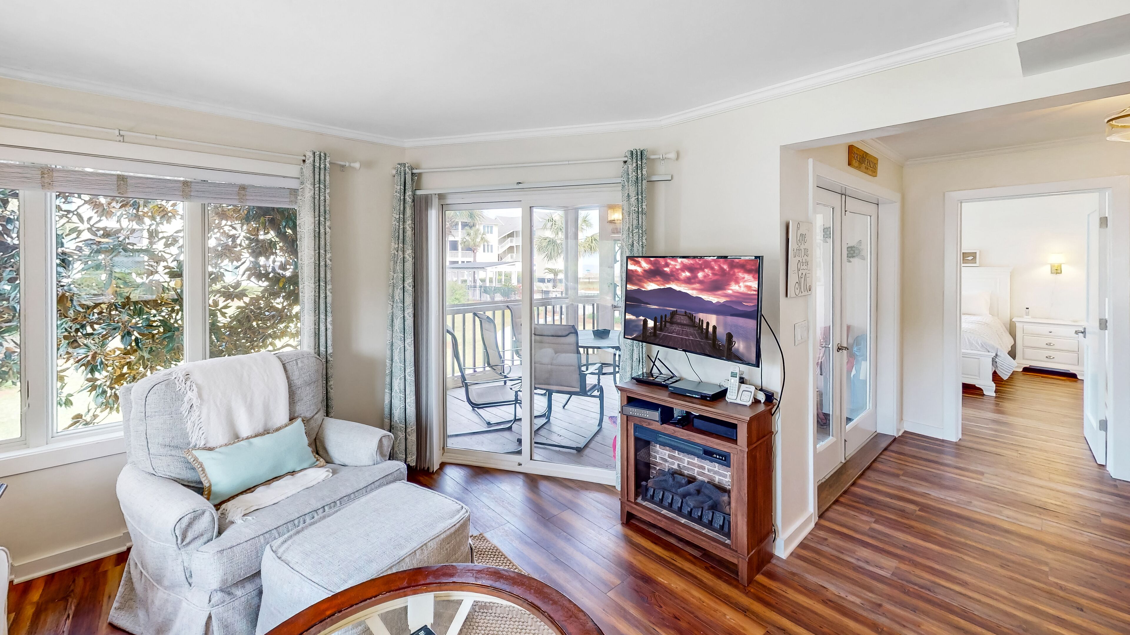 Een woonkamer met een bank, een televisie en een open haard. Er is een balkon met uitzicht op palmbomen.