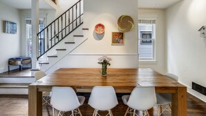 A dining area with a wooden table, three white chairs, a vase with flowers, and a staircase with a railing.