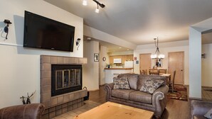 A living room with a fireplace, a television mounted on the wall, and a dining area in the background. - Affordable Purgatory Ski in/Ski Out Condo in the Main Plaza (Durango)