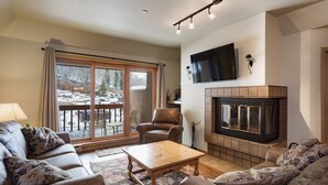 A cozy living room with a fireplace, leather sofas, a wooden coffee table, and a large window with a view of a snowy landscape. - Affordable Purgatory Ski in/Ski Out Condo in the Main Plaza (Durango)