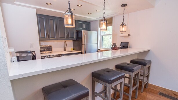 Una cocina moderna con encimera blanca, armarios oscuros, electrodomésticos de acero inoxidable y tres taburetes de bar.