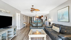 A living room with a flat-screen TV, a ceiling fan, a coffee table, a sofa, and a dining area in the background. - Beachfront - Ocean Views - Beach Place #108 (Madeira Beach)