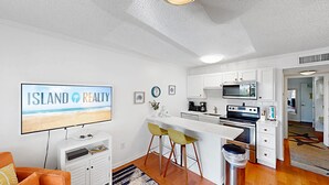 A modern kitchen with white cabinetry, a microwave, stove, and a sink. There's a dining area with a white table and yellow chairs. A TV on the wall displays 'ISLAND REALTY'. - Nestled Just Steps Away from the Sun-Kissed Shores! (Isle of Palms)