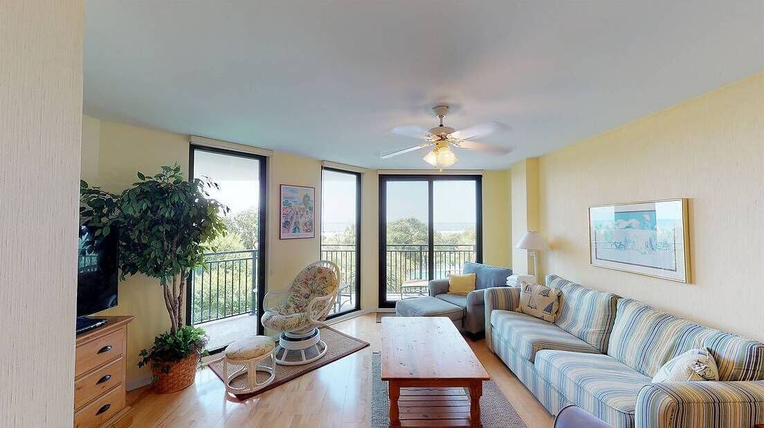 A spacious living room with a large window, a ceiling fan, a sofa set, a coffee table, and a potted plant.