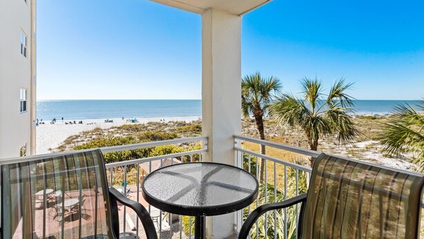 Ein Balkon mit Glastisch und Stühlen, der Blick auf einen Strand mit Palmen und einen klaren, blauen Himmel bietet.